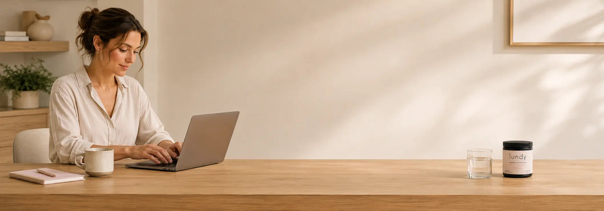 A woman sits at a wooden desk using a laptop in a bright, modern room. A white mug, a notebook, a small glass, and a lundy jar with a white label sit on the desk.