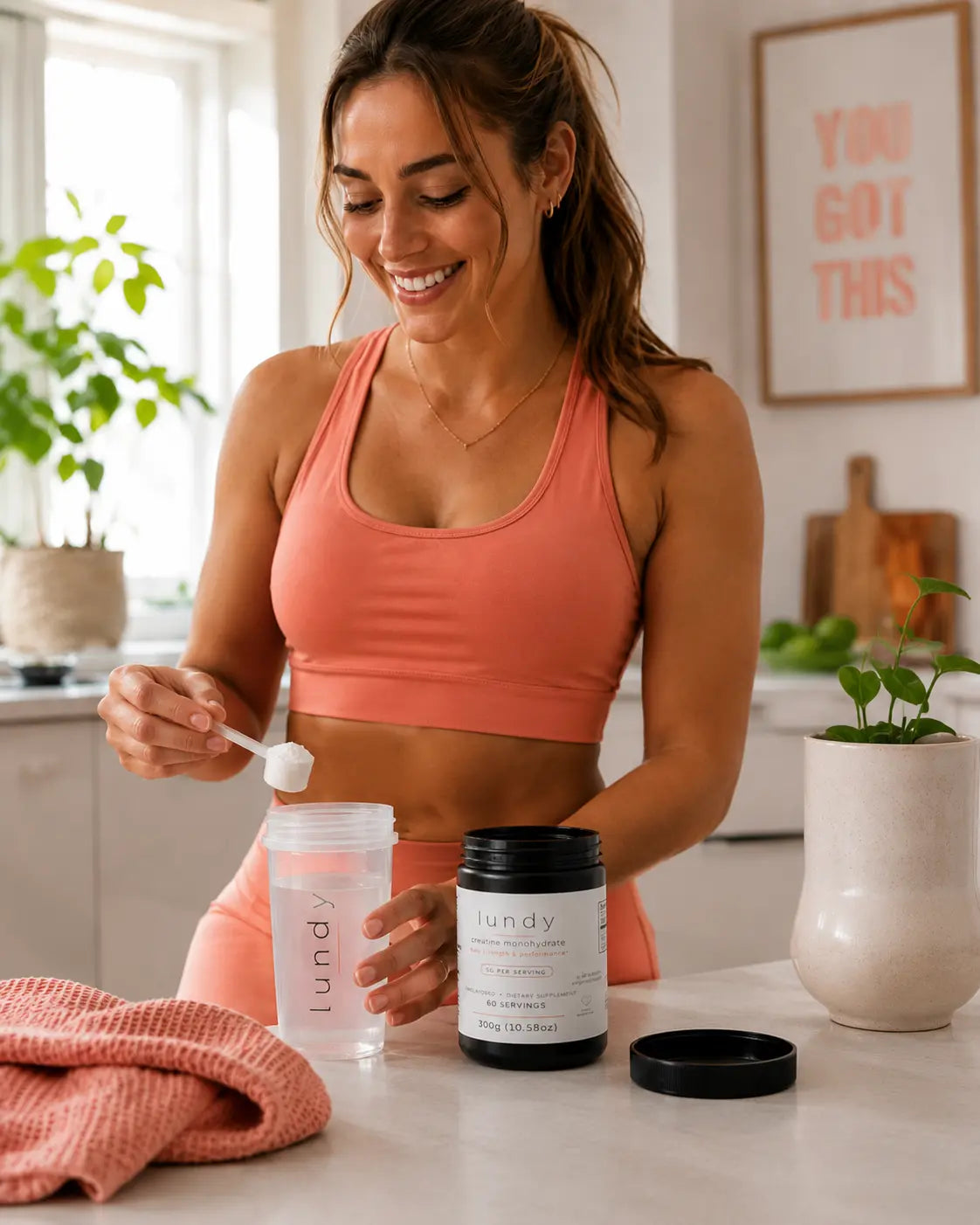 A woman in a coral sports bra stands in a bright kitchen, mixing powder with a scoop and pouring it into a clear bottle. A black tub labeled 'lundy' sits on the counter beside her.