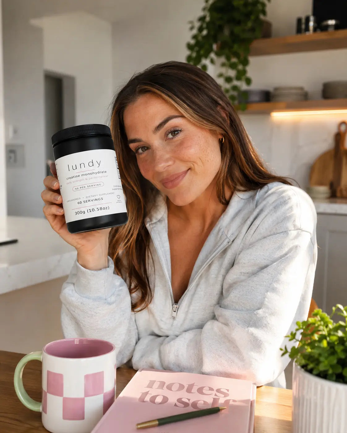 A woman in a light gray zip-up hoodie holds up a black tub of 'lundy' laundry protein powder in a bright kitchen setting. A pink and white checkered mug, a pink notebook, and a small potted plant sit on the table in front of her.