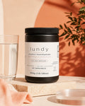 A black tub of lundy creatine monohydrate dietary supplement with a white label, styled on a stone block next to a glass of water with a peach fabric and warm background.