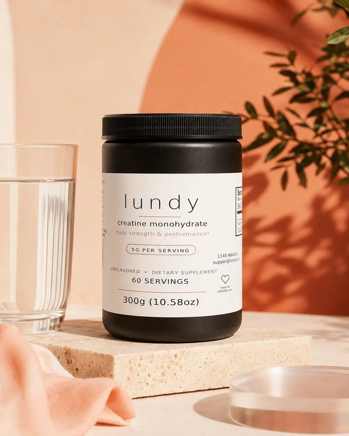 A black tub of lundy creatine monohydrate dietary supplement with a white label, styled on a stone block next to a glass of water with a peach fabric and warm background.