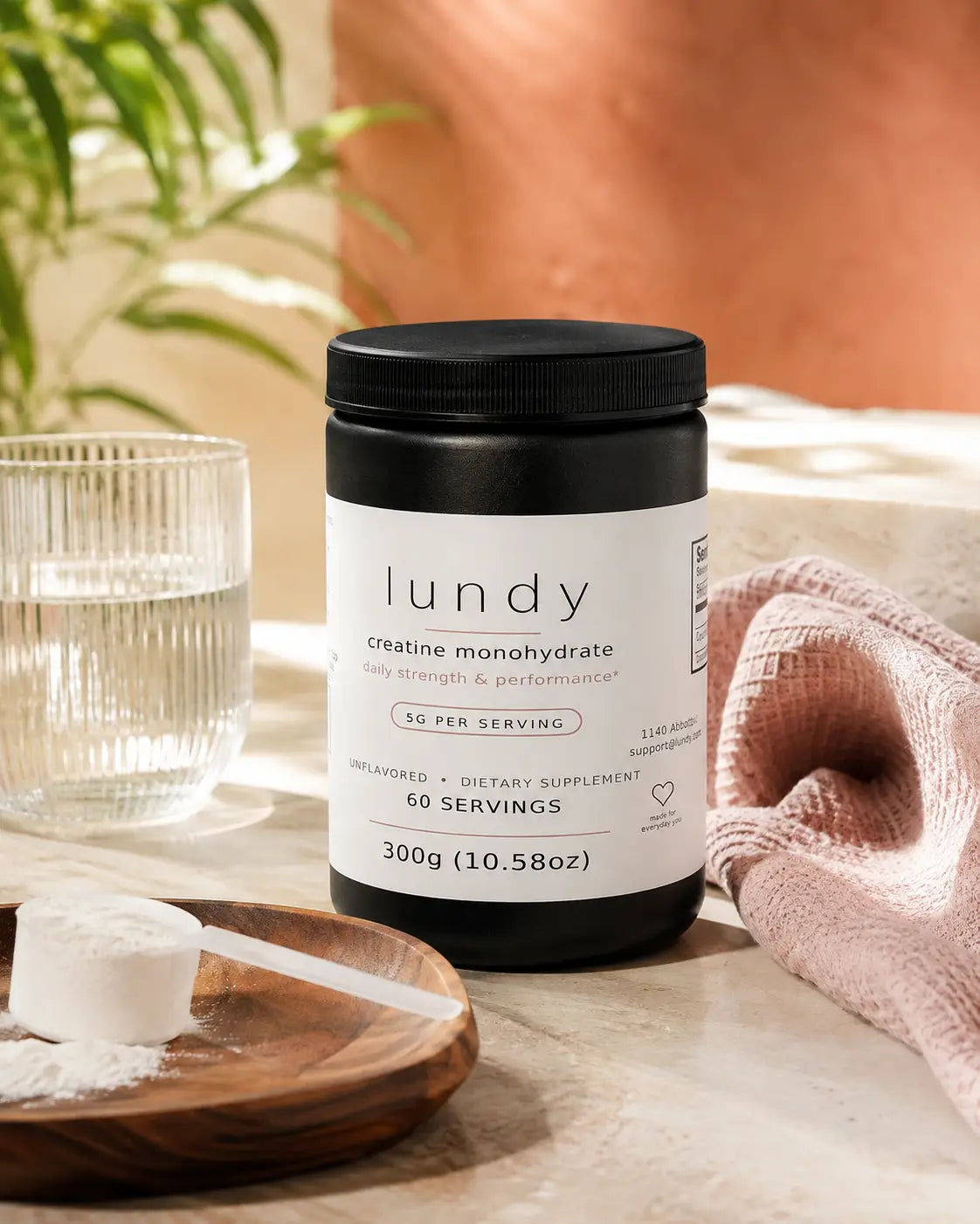 A black tub of lundy creatine monohydrate dietary supplement with a white label, styled on a marble surface with a glass of water, a wooden scoop of white powder, and a pink textured cloth.