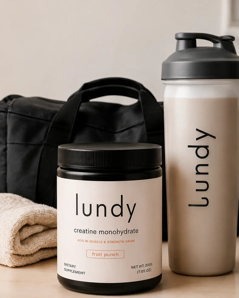 A lifestyle product shot featuring a black supplement tub of 'lundy creatine monohydrate' next to a matching beige shaker bottle with a dark gray lid, both branded with the 'lundy' logo. A black duffel bag and a beige towel are visible in the background on a neutral beige surface.