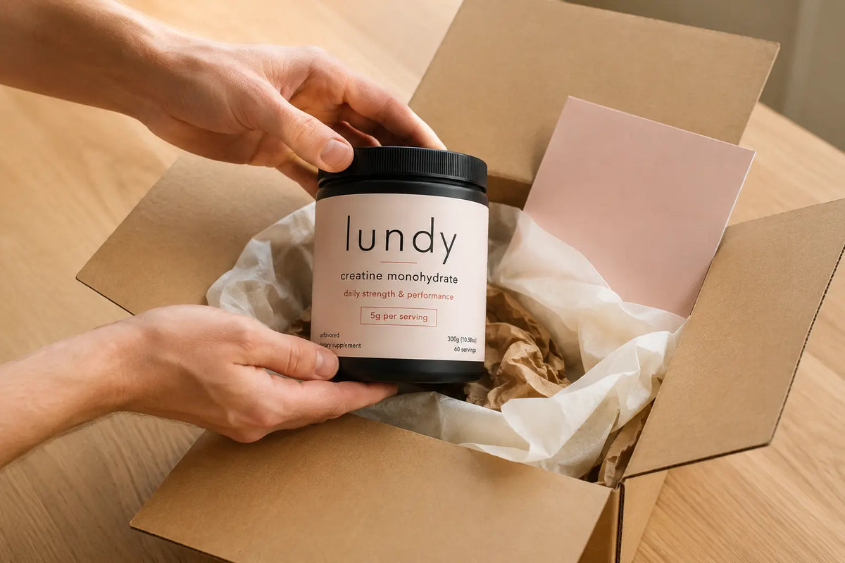 A person holds a black tub of lundy creatine monohydrate over an open brown cardboard gift box with tissue paper on a wooden table.