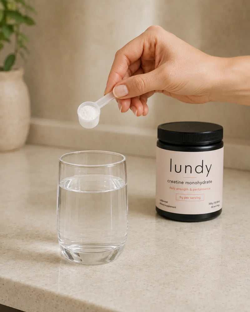 A hand holds a small white scoop above a glass of water, with a black jar labeled 'lundy creatine monohydrate' standing beside it on a light countertop.