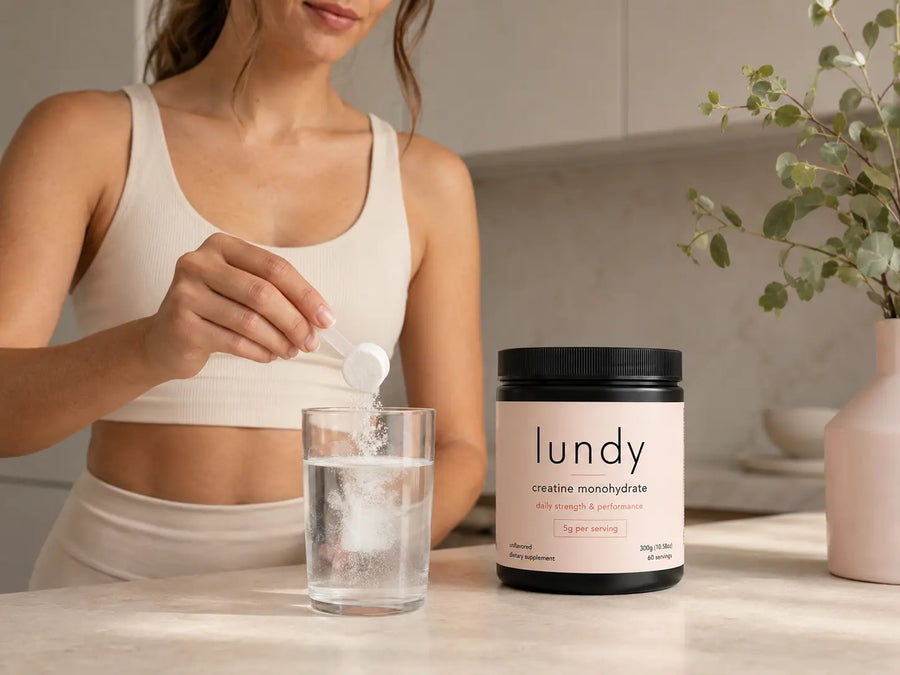 A woman in a beige sports bra pours powder from a white scoop into a glass of water, with a black and cream jar of lundy creative monohydrate on the counter next to her.